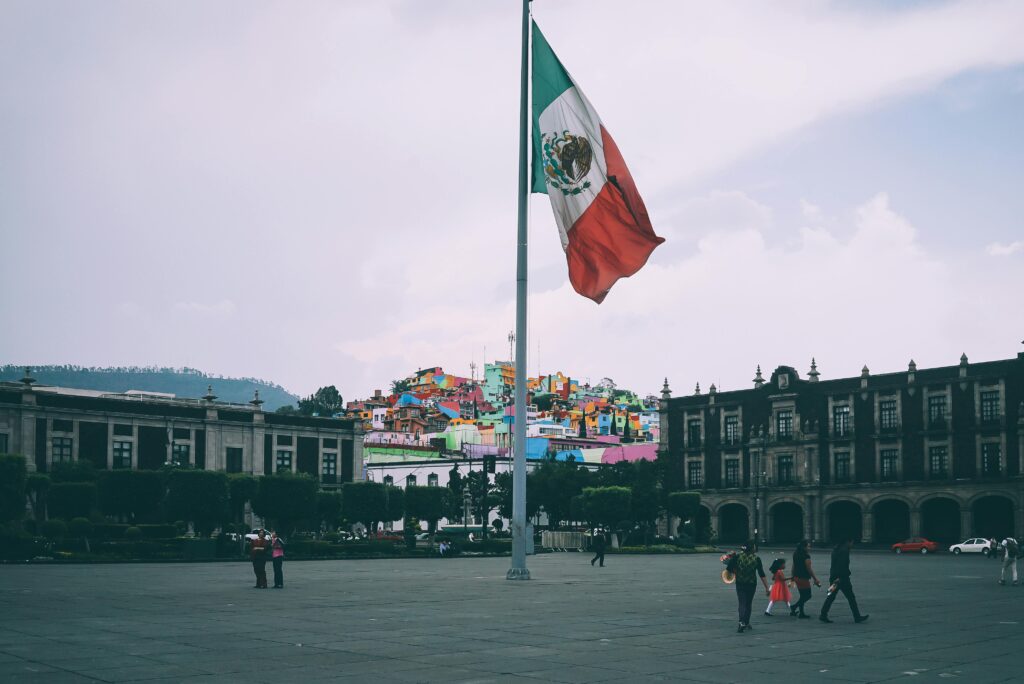 Mexico: The Tanda System Colorful cityscape of Toluca with large Mexican flag in the foreground, vibrant buildings in the backdrop.