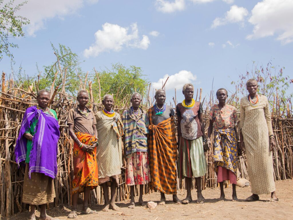 Kenya: Harambee (Community Saving) Group of senior adults in colorful traditional clothing standing outdoors in a village setting.